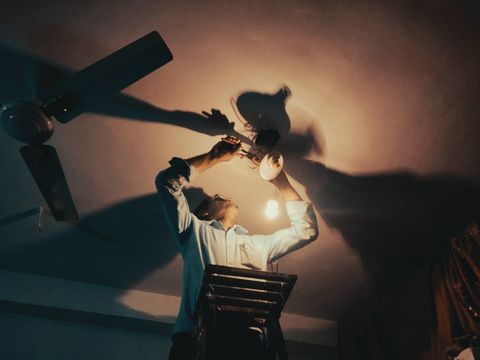 An electrician on a ladder fitting a ceiling fan, lit from below